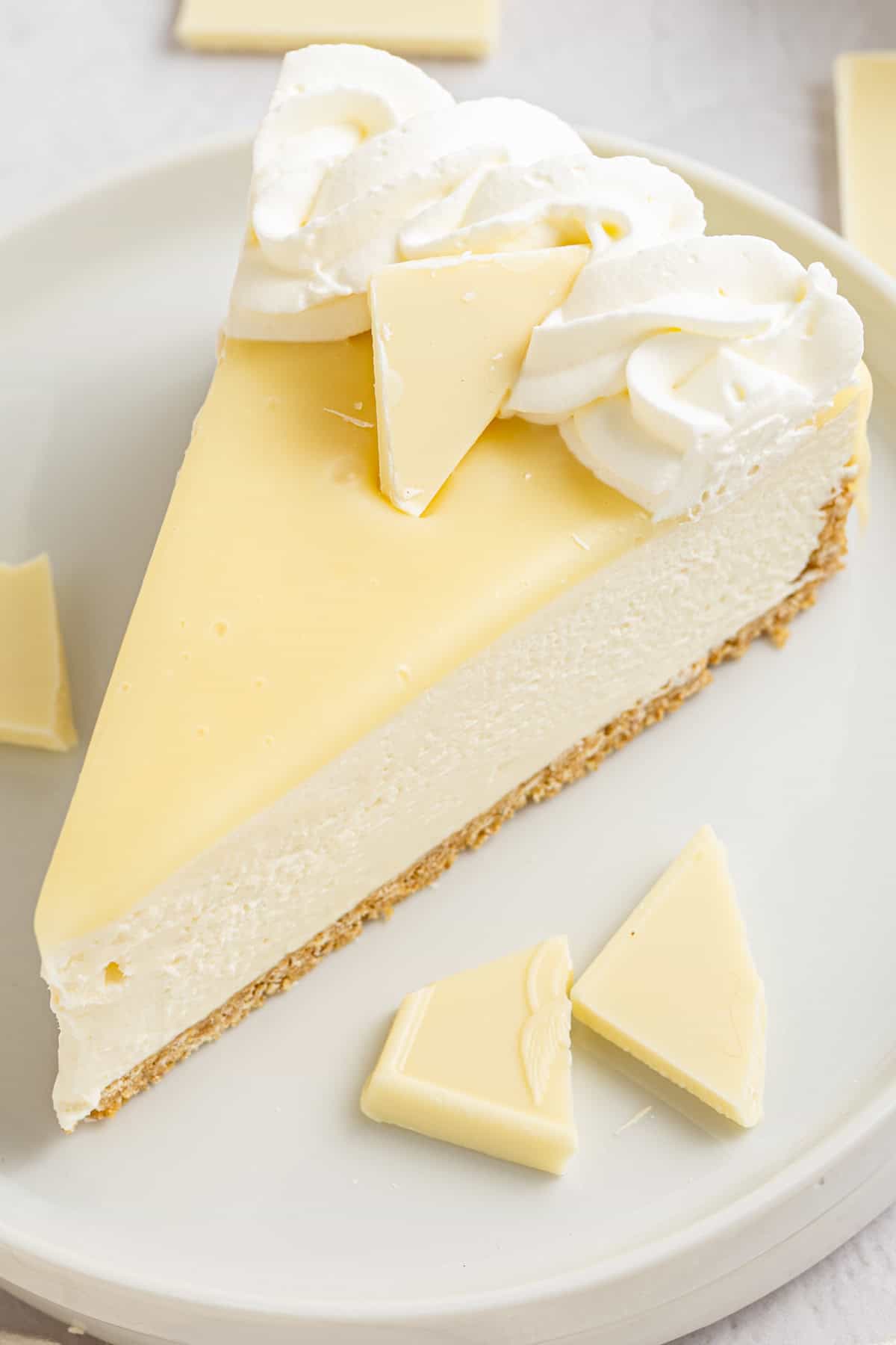White Chocolate Cheesecake A slice of white chocolate cheesecake with a graham cracker crust, topped with whipped cream and pieces of white chocolate.