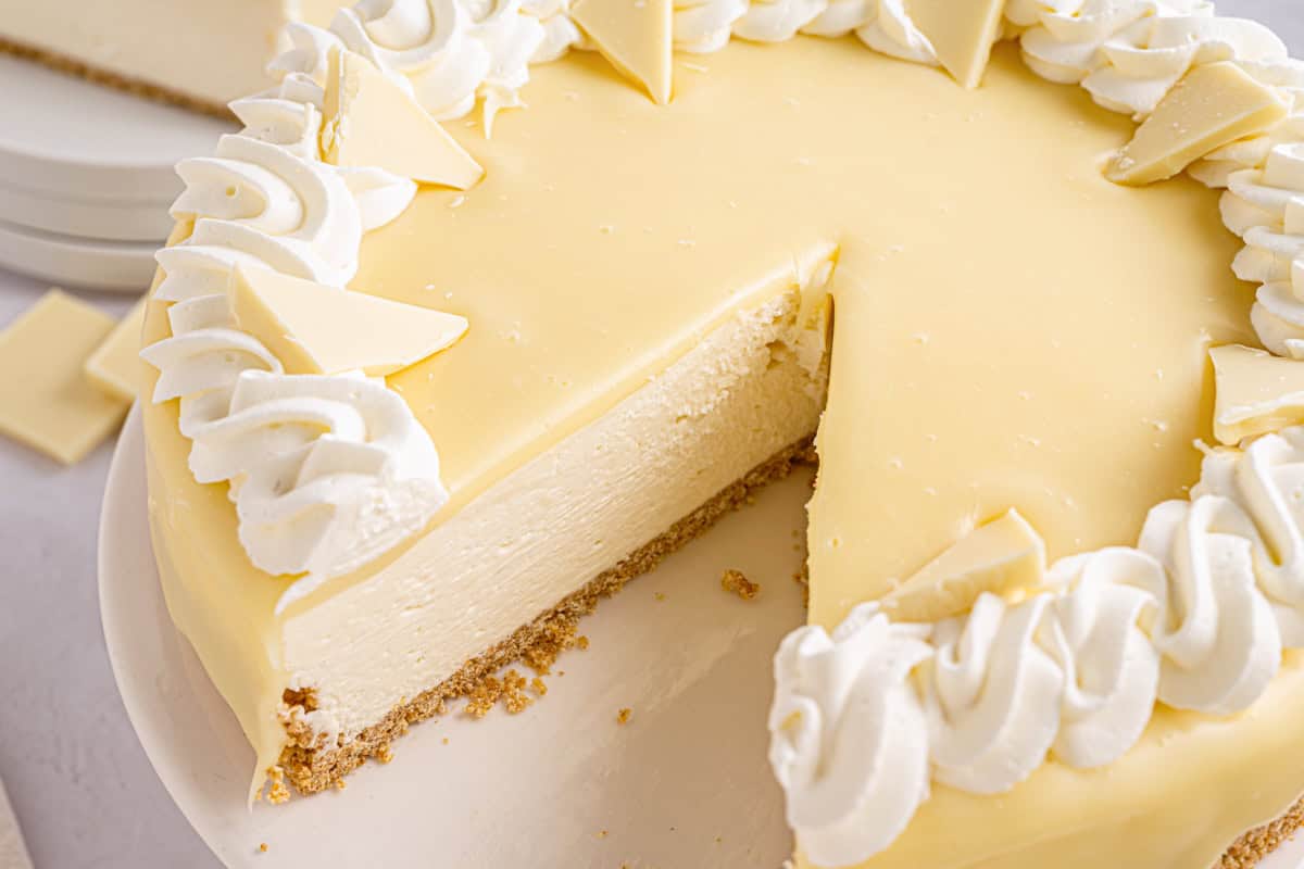 White Chocolate Cheesecake A creamy white cheesecake with a slice removed, topped with white chocolate pieces and whipped cream.
