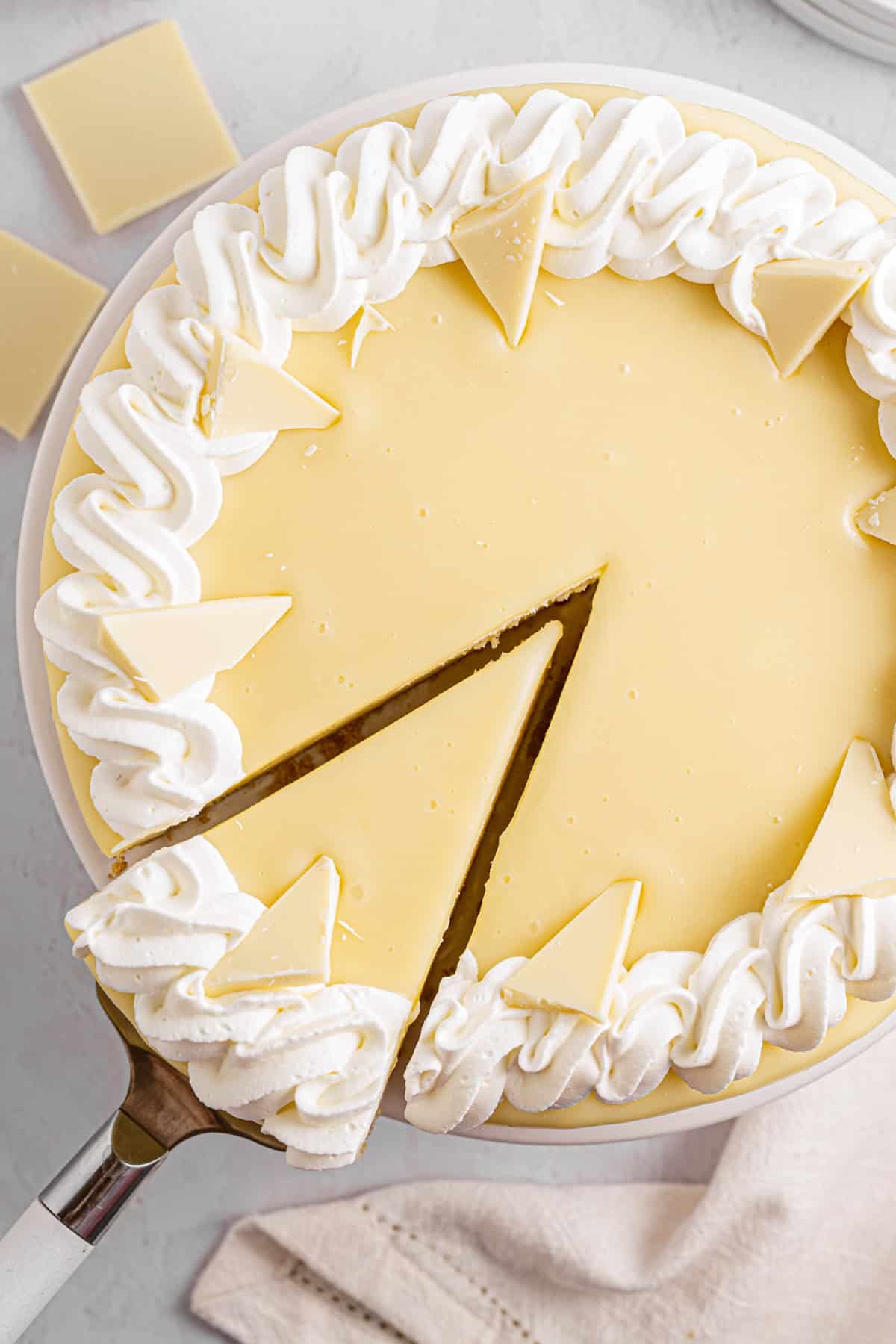 White Chocolate Cheesecake A white chocolate cheesecake is topped with whipped cream and white chocolate pieces. One slice is being lifted from the cake with a pie server.
