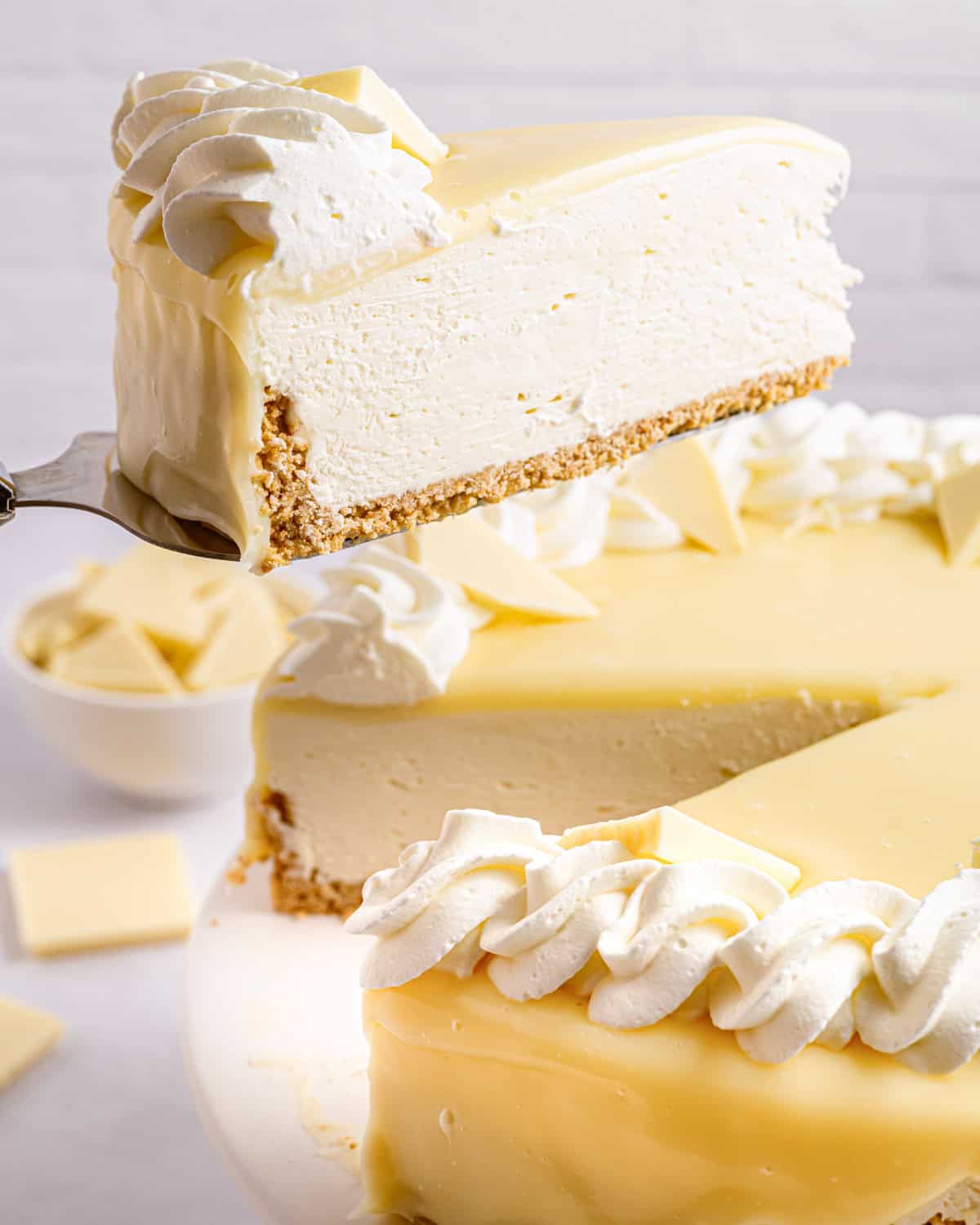 White Chocolate Cheesecake A slice of creamy white cheesecake with a graham crust and whipped cream is being lifted from a whole cheesecake, which is topped with white chocolate pieces and swirls of whipped cream.