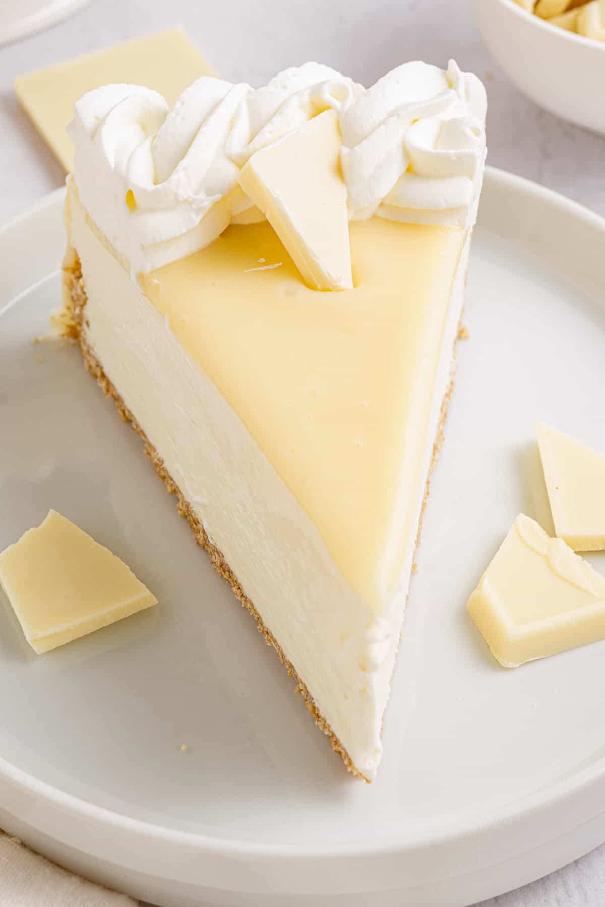 White Chocolate Cheesecake A slice of white chocolate cheesecake topped with whipped cream and a piece of white chocolate on a white plate.