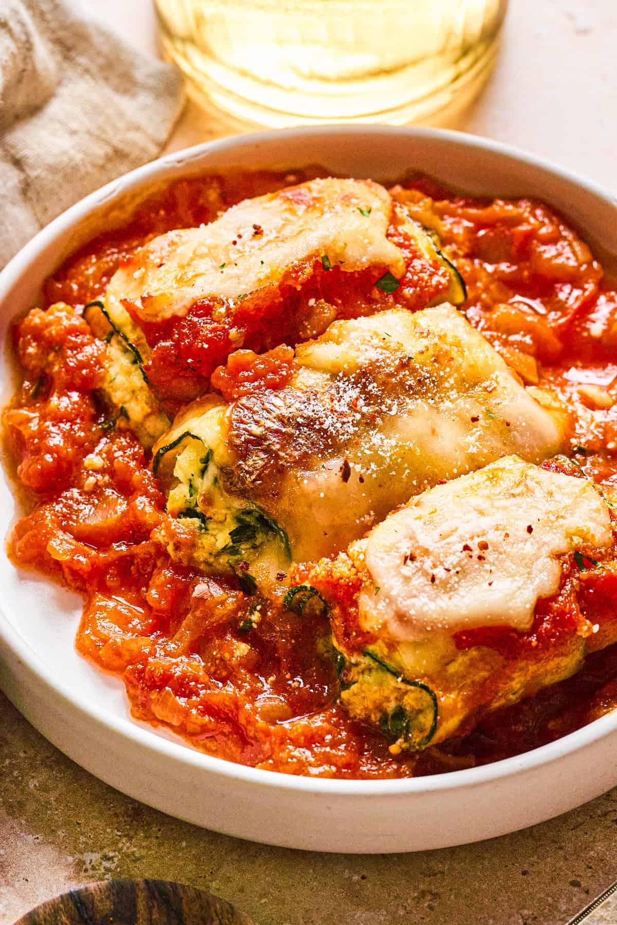 Zucchini Rollatini Zucchini roll-ups filled with cheese and herbs, sit on chunky tomato sauce in a white bowl.