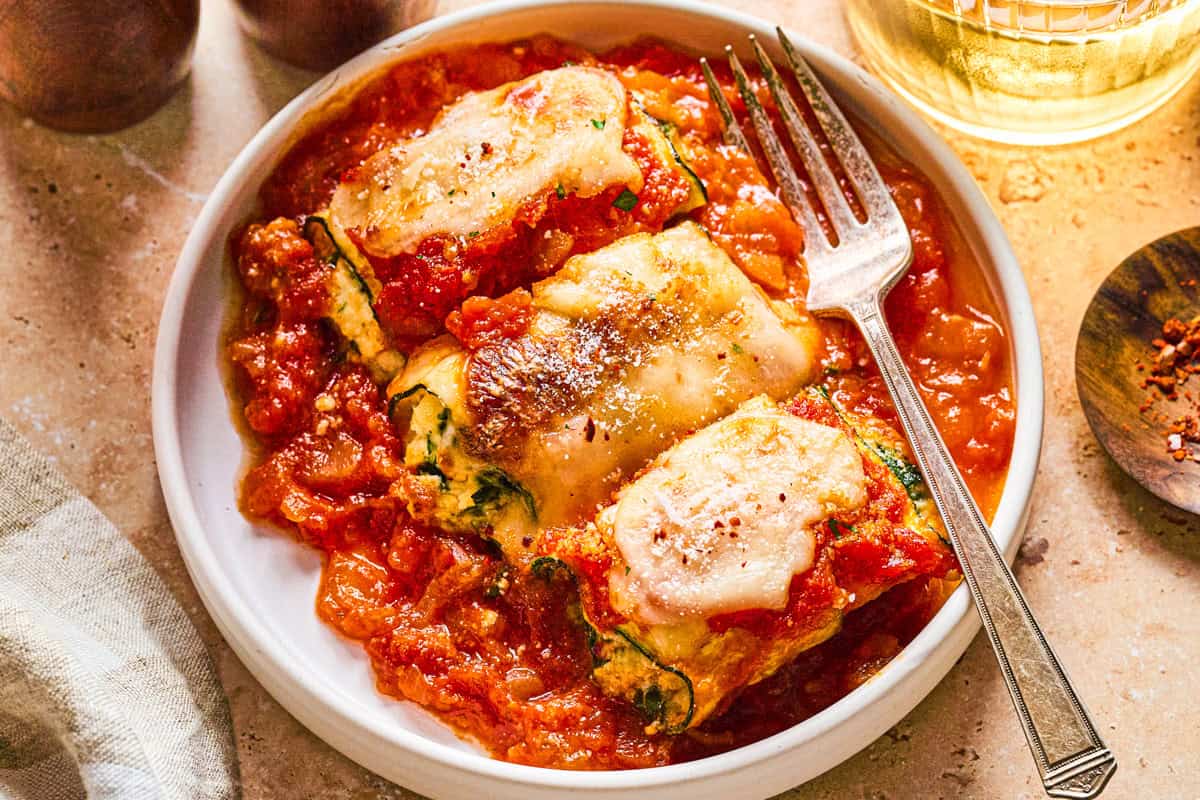Zucchini Rollatini A plate with three zucchini rolls stuffed with cheese and topped with tomato sauce and melted cheese, served with a fork on the side.