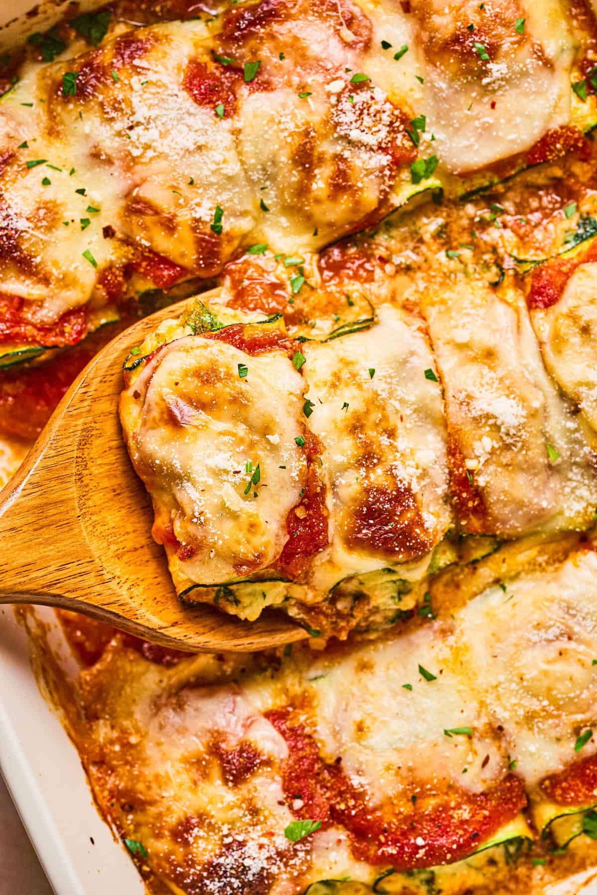 Zucchini Rollatini A wooden spatula lifts a cheesy, baked zucchini slice from a casserole dish, showing layers of sauce, melted cheese, and herbs on top.