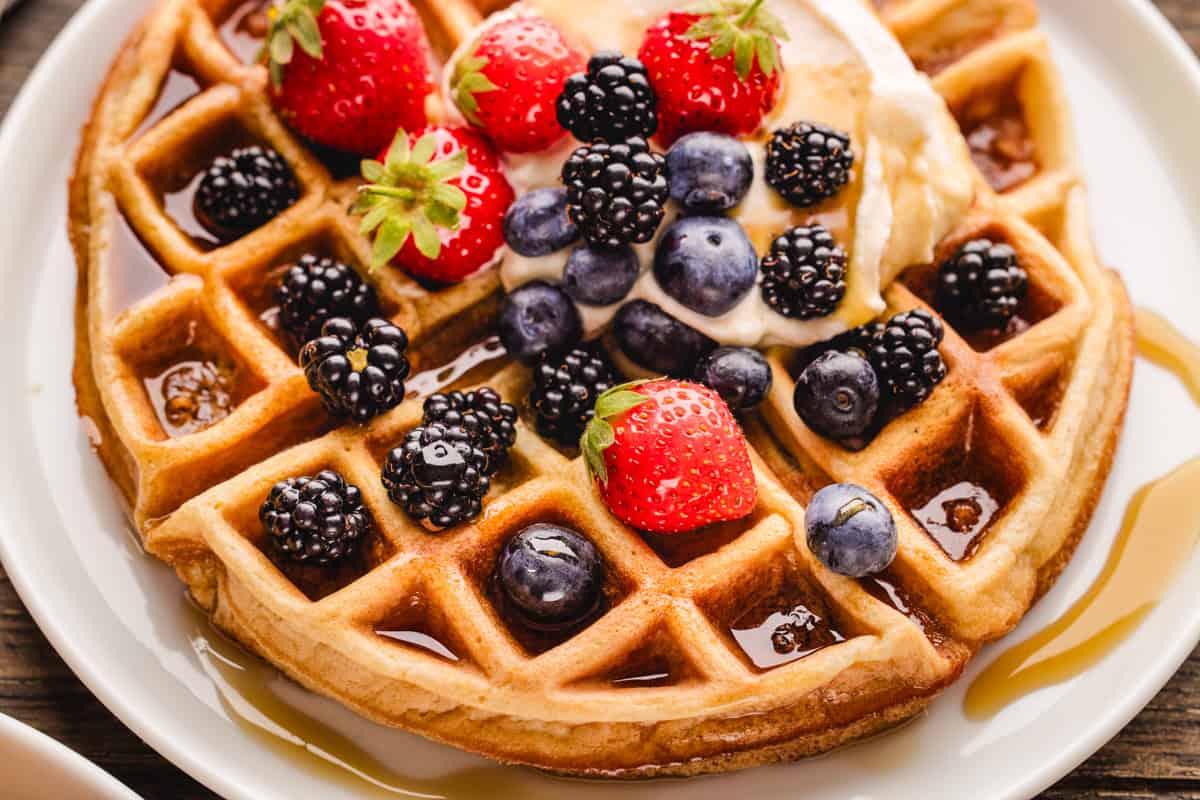 A close-up of a golden waffle topped with strawberries, blackberries, blueberries, cream, and syrup on a white plate.