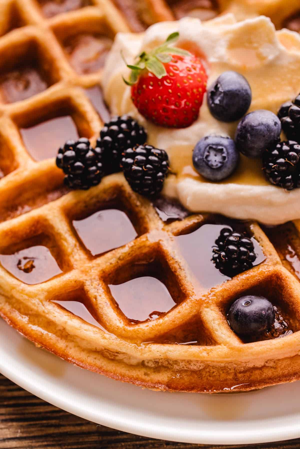 Close-up of a golden waffle topped with blackberries, blueberries, a strawberry, cream, and syrup on a white plate.