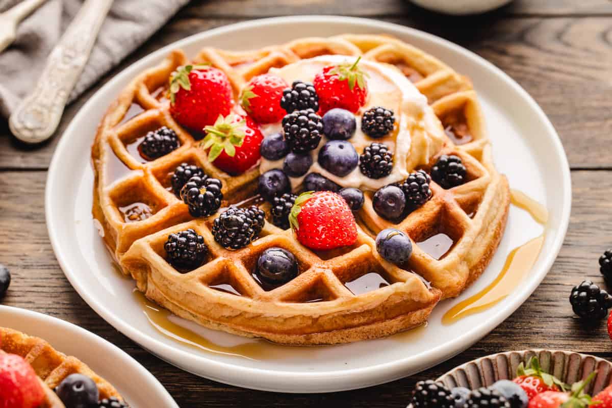 A waffle topped with cream, strawberries, blueberries, and blackberries sits on a white plate, drizzled with syrup on a wooden table.