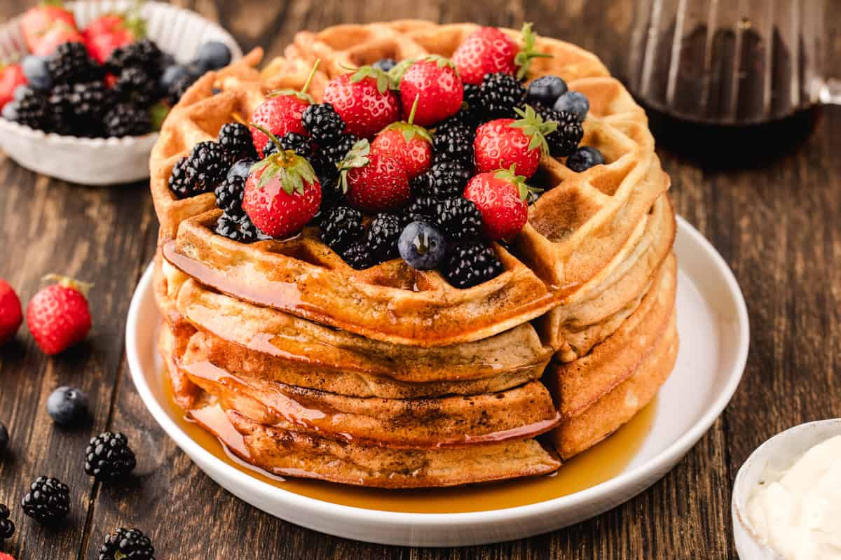 A stack of waffles topped with fresh strawberries, blackberries, and blueberries, drizzled with syrup, sits on a white plate on a wooden table.