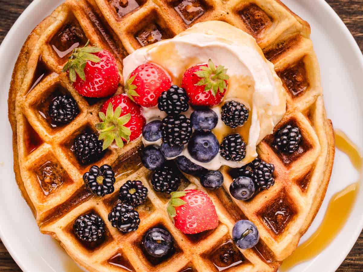 A plate of golden waffles topped with whipped cream, strawberries, blueberries, blackberries, and a drizzle of syrup.