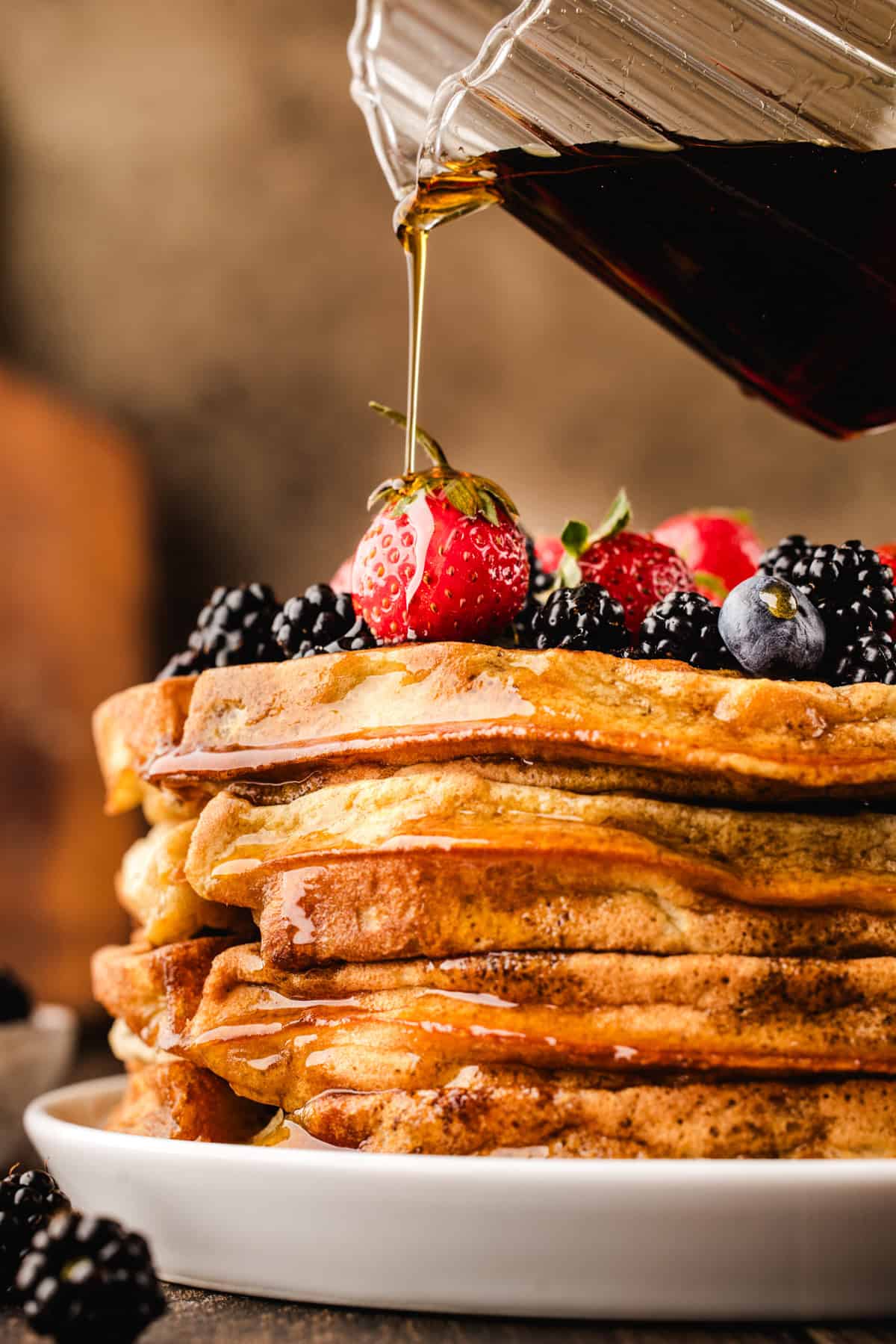 A stack of pancakes topped with strawberries, blackberries, and blueberries is being drizzled with syrup.
