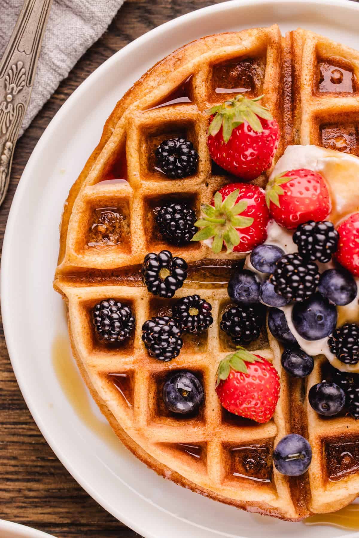 A golden waffle topped with strawberries, blackberries, blueberries, and cream sits on a white plate with syrup pooling in the squares. 