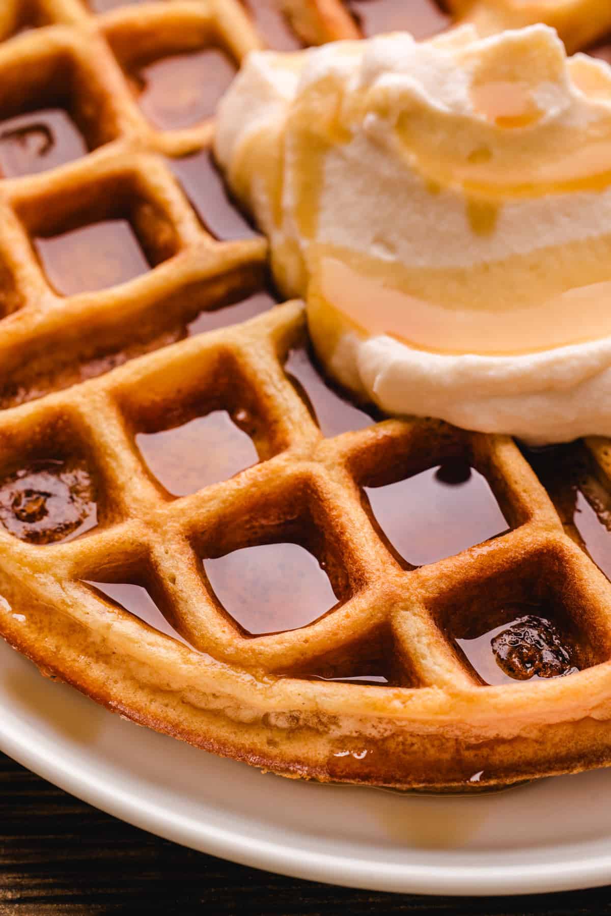 Close-up of a golden waffle topped with cream and drizzled with syrup, served on a white plate.