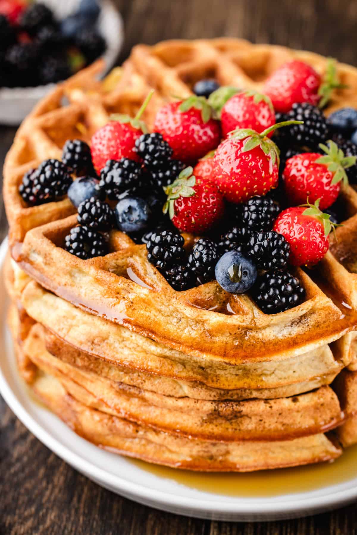 A stack of golden waffles topped with fresh strawberries, blueberries, and blackberries sits on a white plate, with syrup drizzling down the sides.