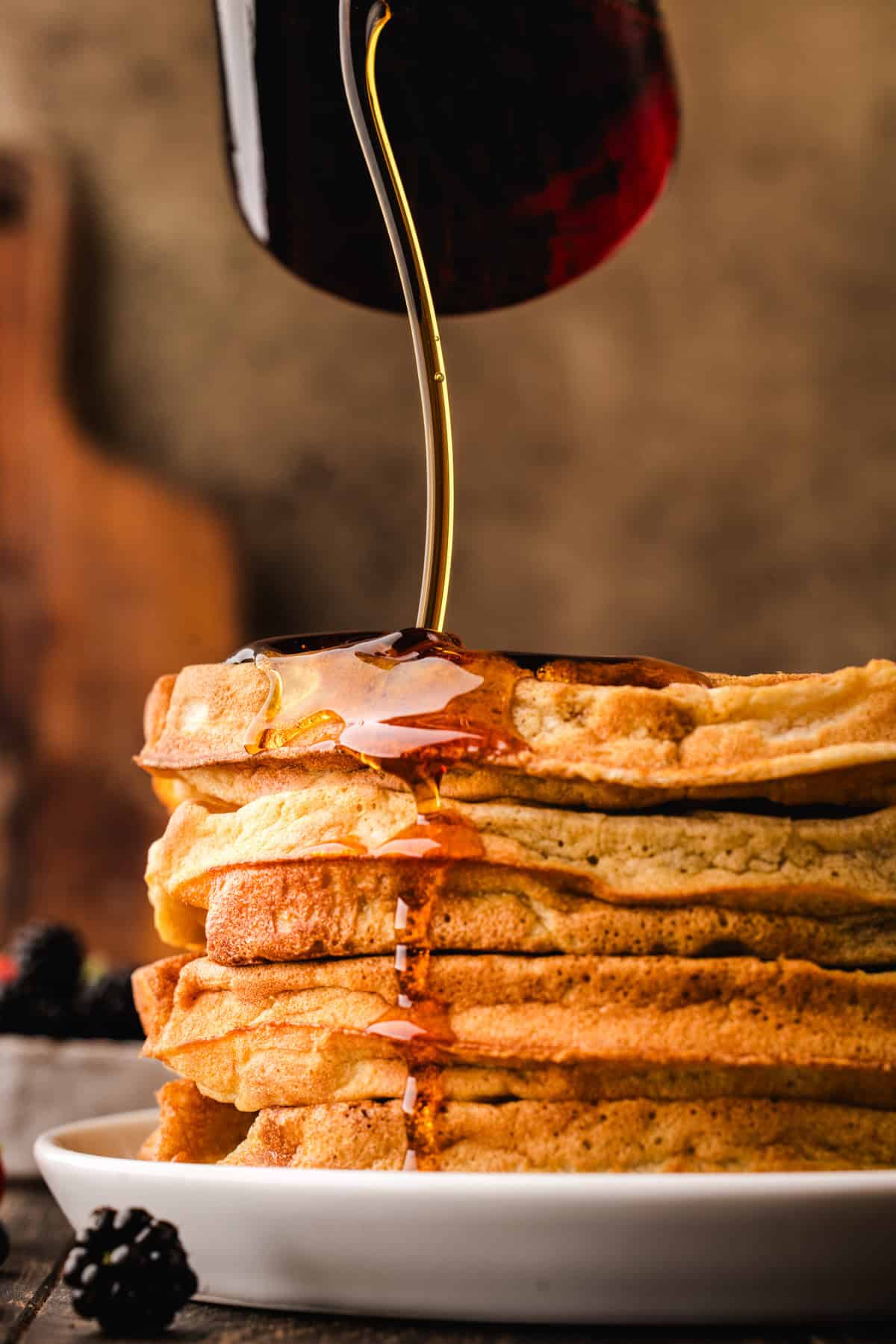 A stack of golden pancakes on a white plate with syrup being poured over them, dripping down the sides.
