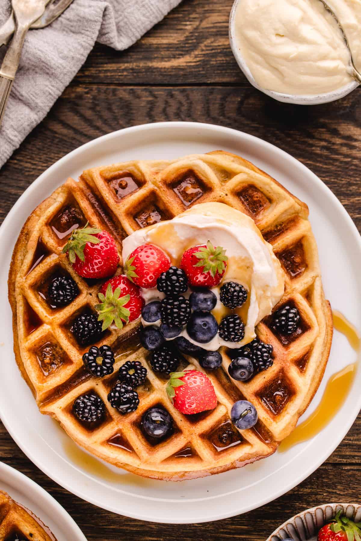 A golden waffle topped with strawberries, blackberries, blueberries, cream, and syrup sits on a white plate on a wooden table.
