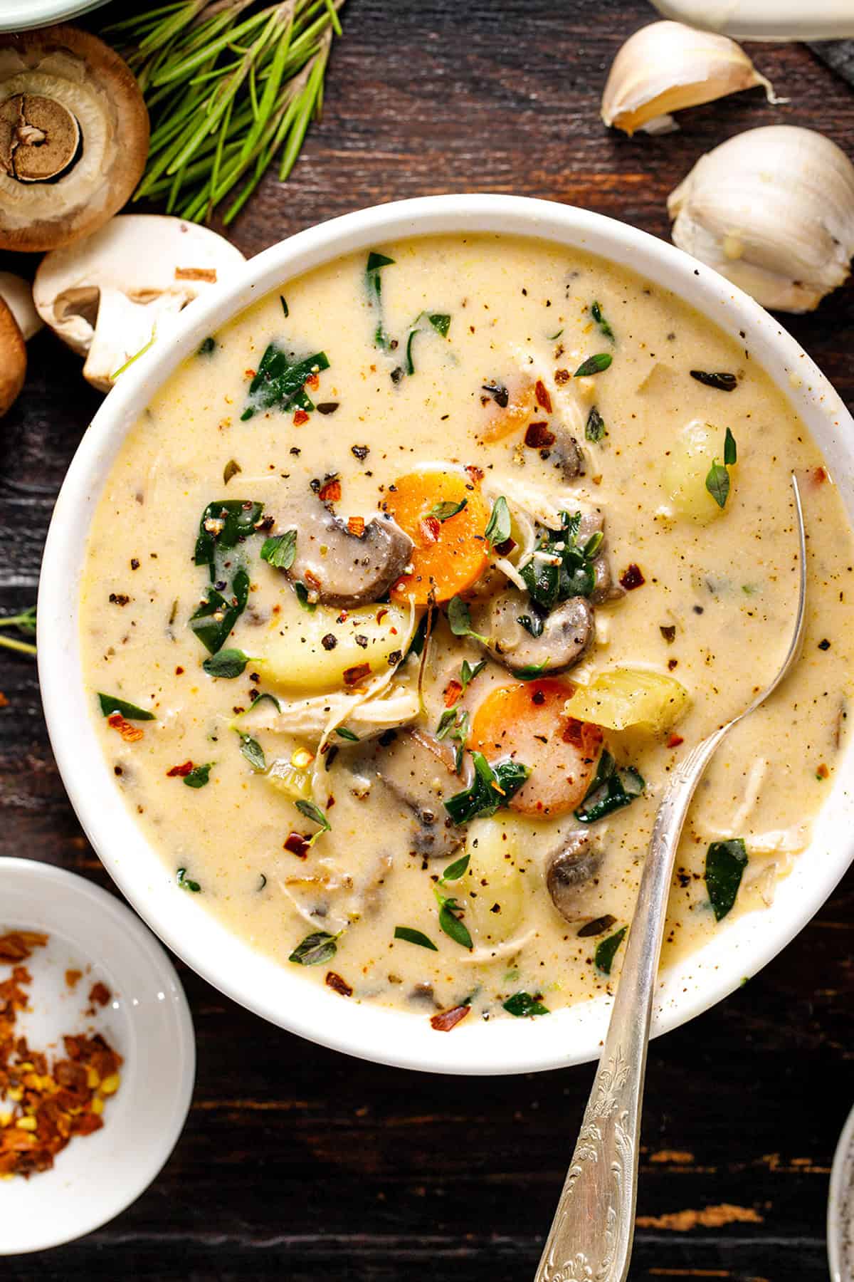 Chicken Gnocchi Soup A bowl of creamy soup with mushrooms, carrots, spinach, and herbs sits on a dark wooden table, garnished with pepper flakes, with a spoon inside.