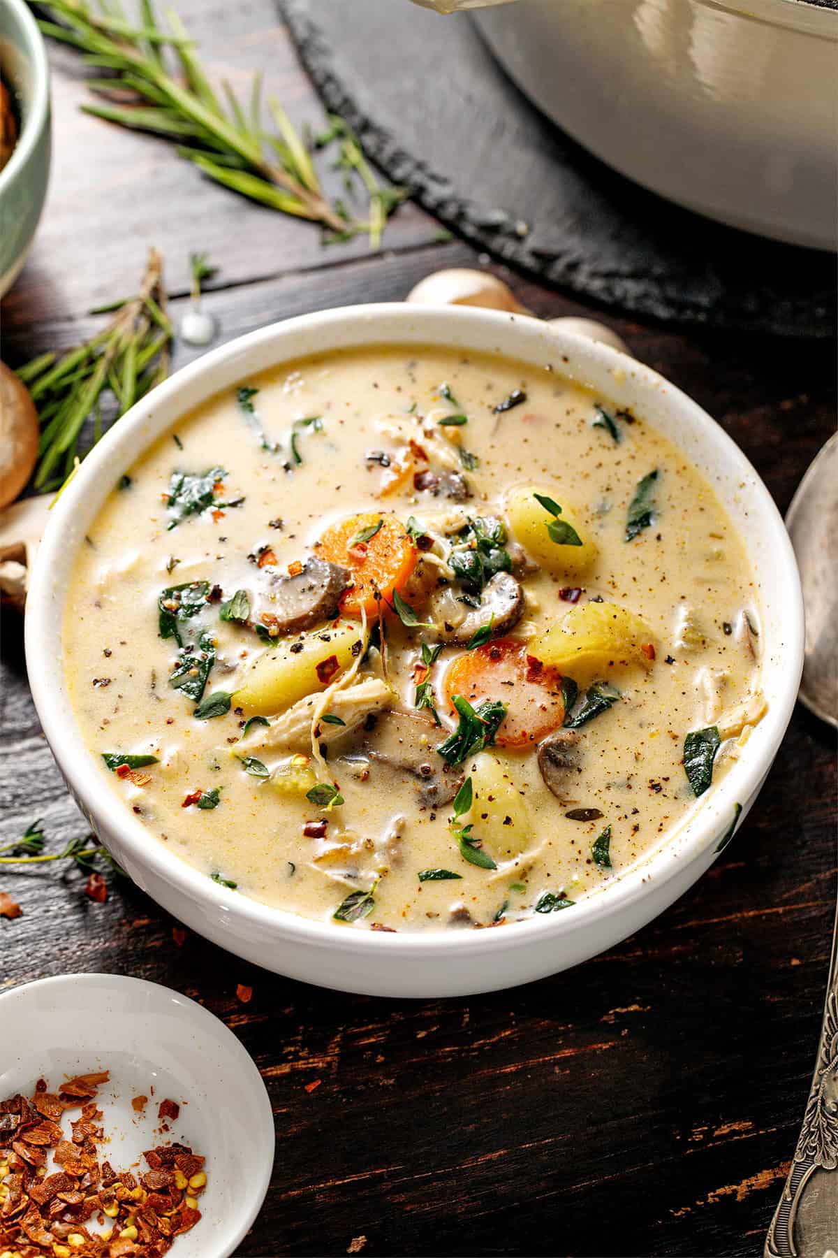 Chicken Gnocchi Soup A creamy soup in a white bowl with visible carrots, potatoes, mushrooms, greens, and herbs, topped with black pepper and chili flakes.