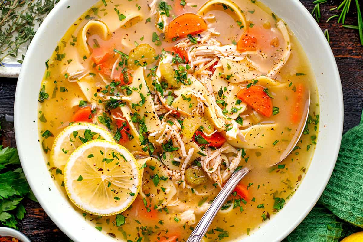 Chicken Noodle Soup A bowl of chicken noodle soup with shredded chicken, carrots, celery, noodles, sliced lemon, fresh herbs, and black pepper, with a spoon inside.