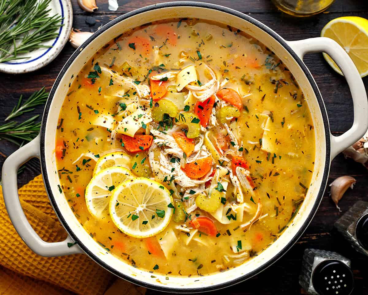 Chicken Noodle Soup A pot of homemade chicken soup with noodles, carrots, celery, herbs, and lemon slices sits on a dark wooden table.