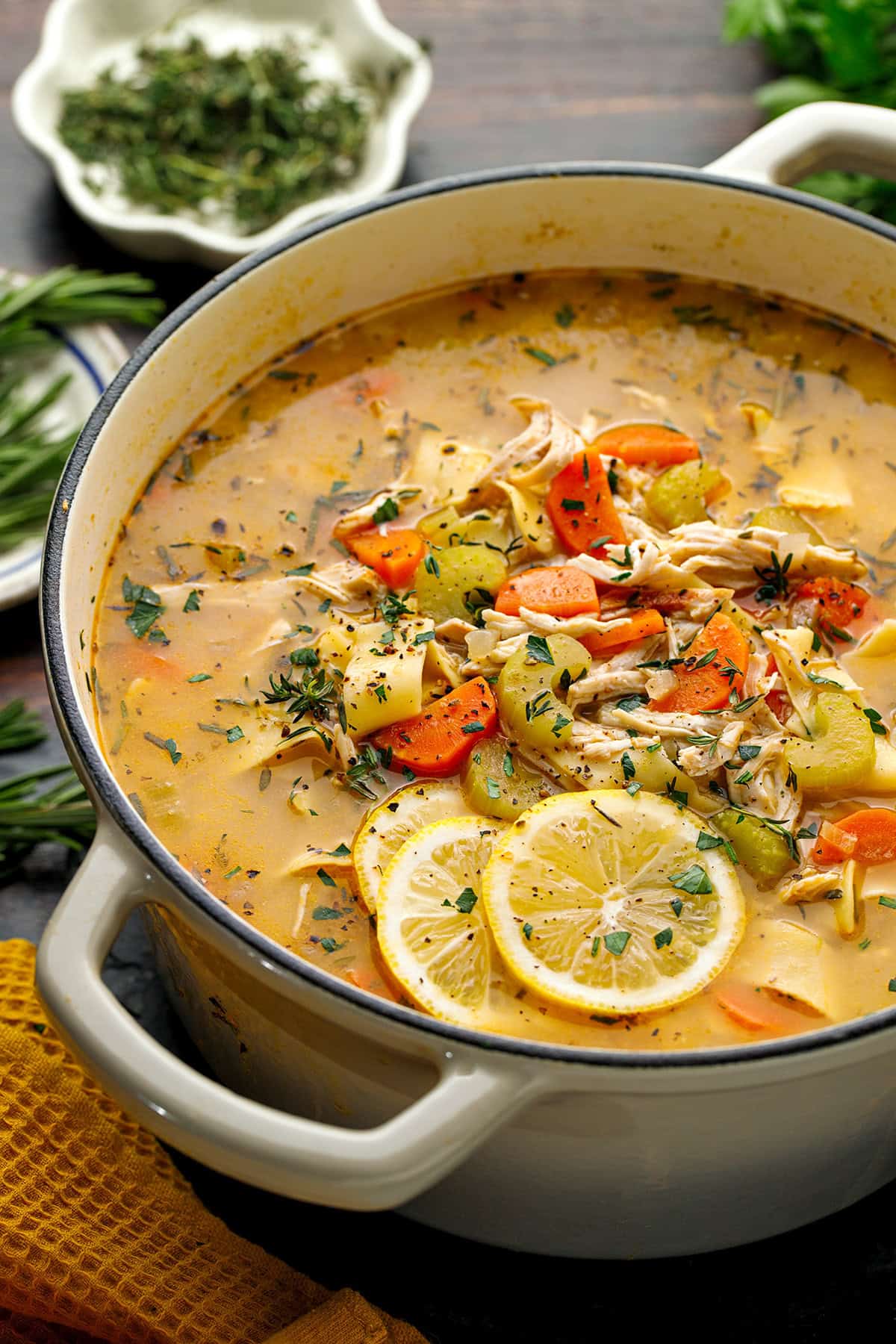 Chicken Noodle Soup A pot of chicken soup with carrots, celery, potatoes, shredded chicken, herbs, and lemon slices, sitting on a table with fresh herbs nearby.