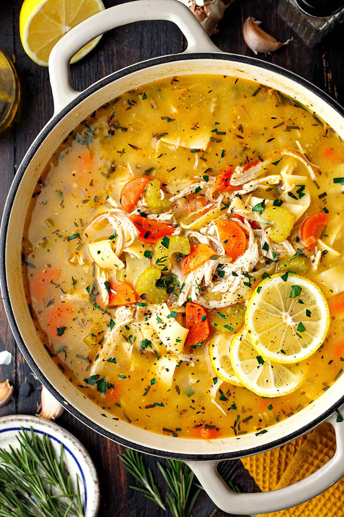 Chicken Noodle Soup A pot of chicken noodle soup with shredded chicken, carrots, celery, herbs, and lemon slices, viewed from above on a dark wooden table.