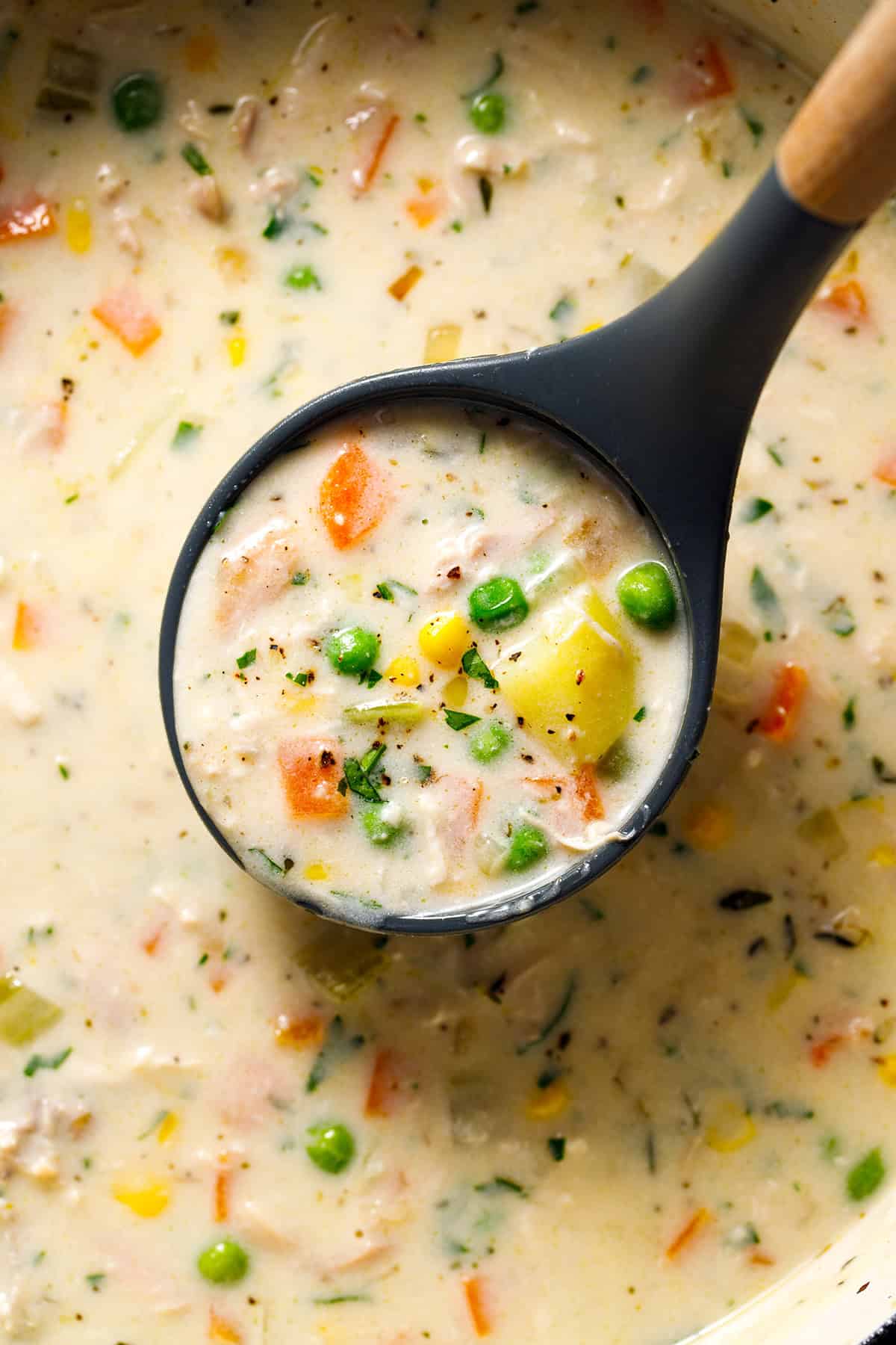 Chicken Pot Pie Soup A black ladle lifts creamy vegetable soup with peas, carrots, corn, potatoes, and herbs from a pot.