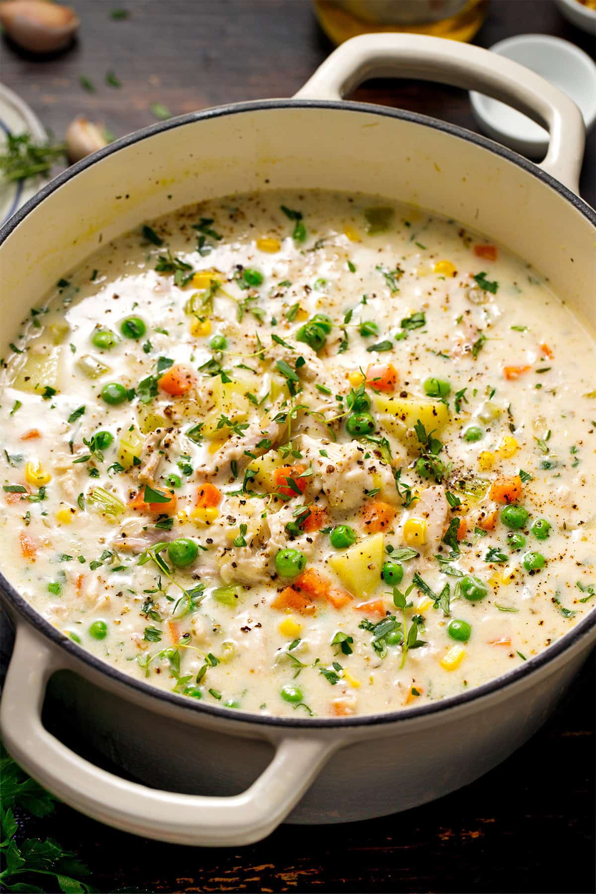 Chicken Pot Pie Soup Creamy vegetable soup with peas, carrots, potatoes, and corn, garnished with herbs and black pepper, simmering in a white pot on a wooden table.