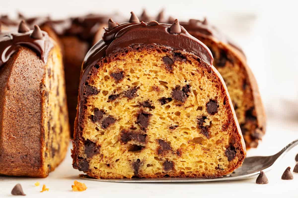 A close-up of a chocolate chip bundt cake slice on a spatula, topped with chocolate frosting and chocolate chips. 