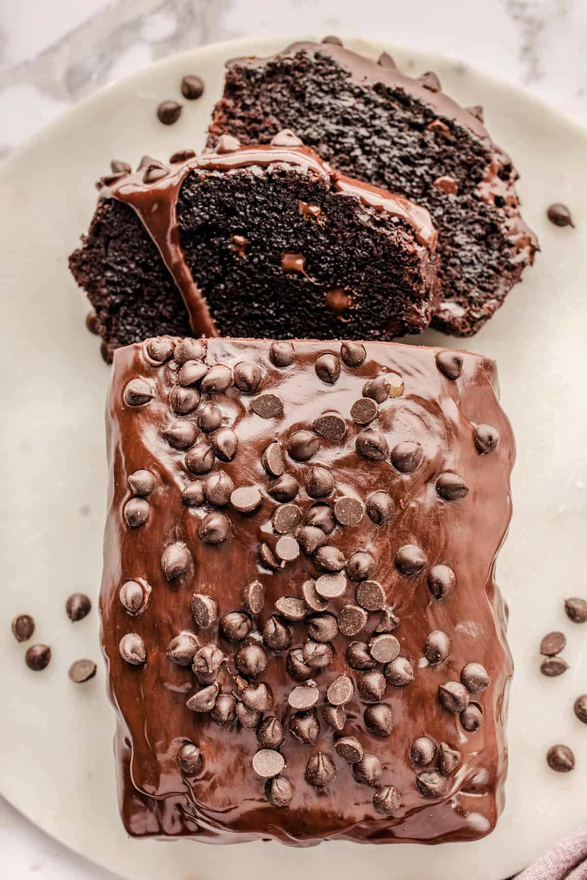 Chocolate Loaf Cake A chocolate loaf cake topped with chocolate chips and glossy chocolate glaze, with two thick slices cut and laid beside the loaf on a white surface.