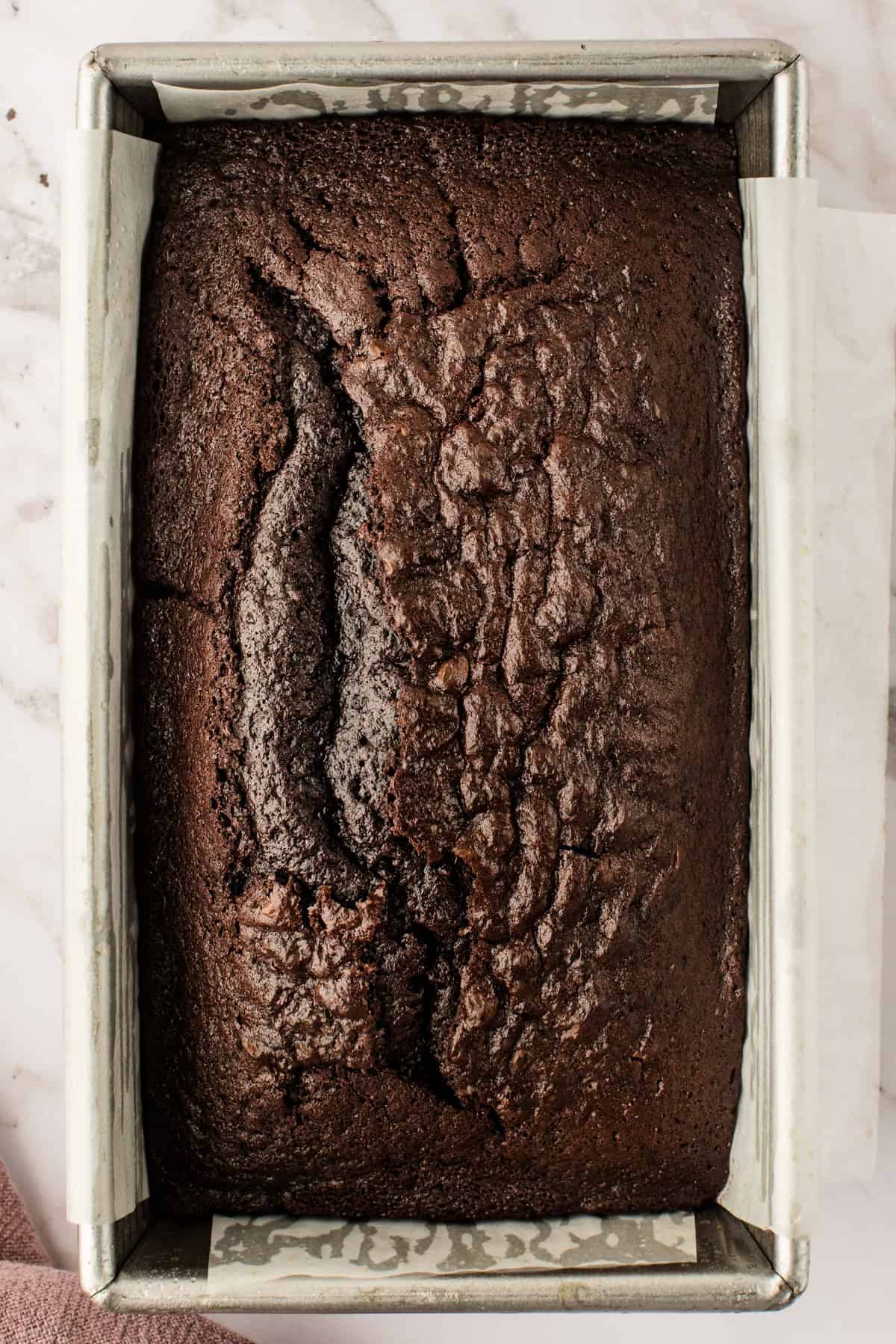 Chocolate Loaf Cake A loaf of chocolate bread with a cracked top sits in a metal baking pan lined with parchment paper, viewed from above.