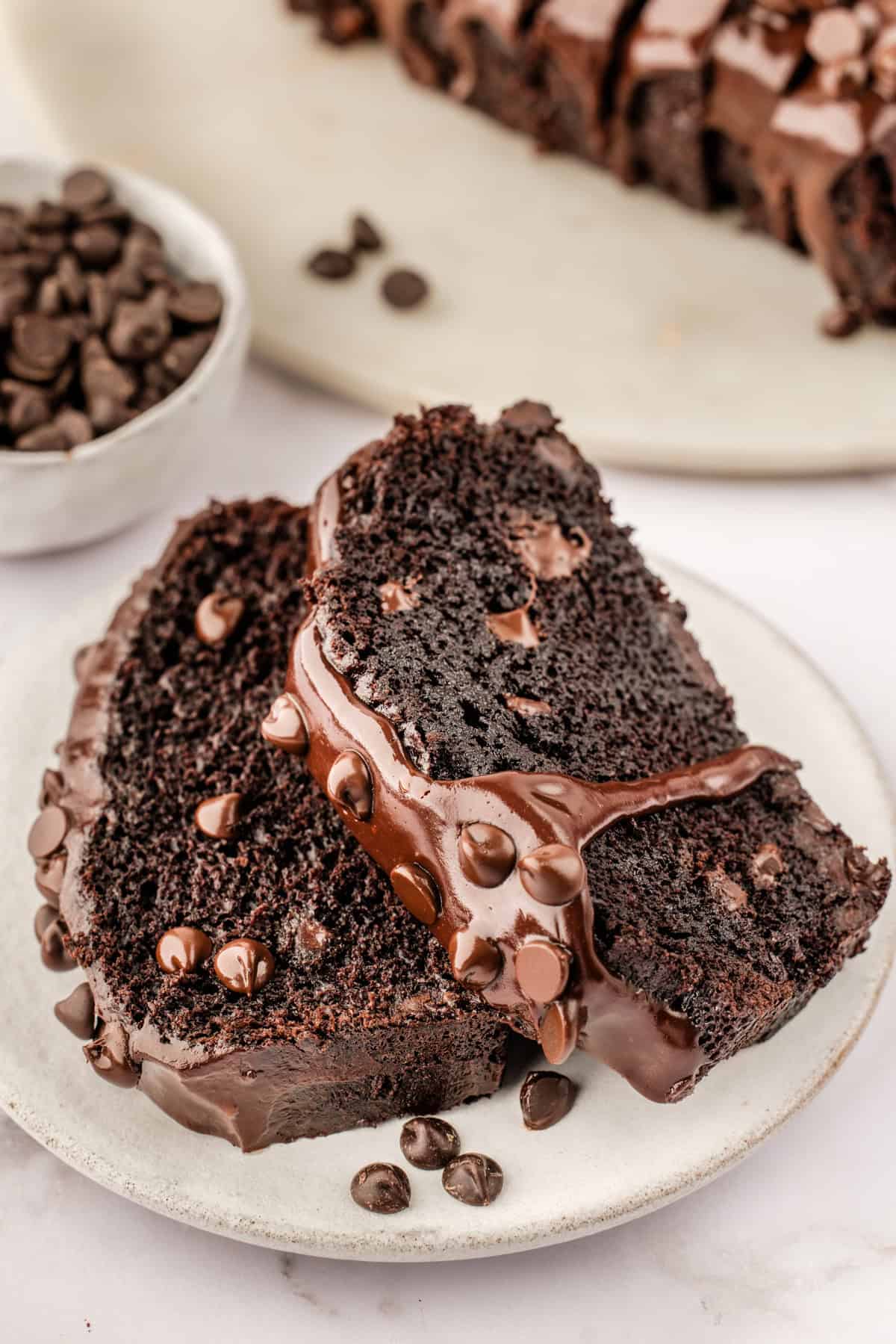 Chocolate Loaf Cake Two slices of rich chocolate loaf cake topped with chocolate chips and glossy chocolate glaze, served on a white plate.