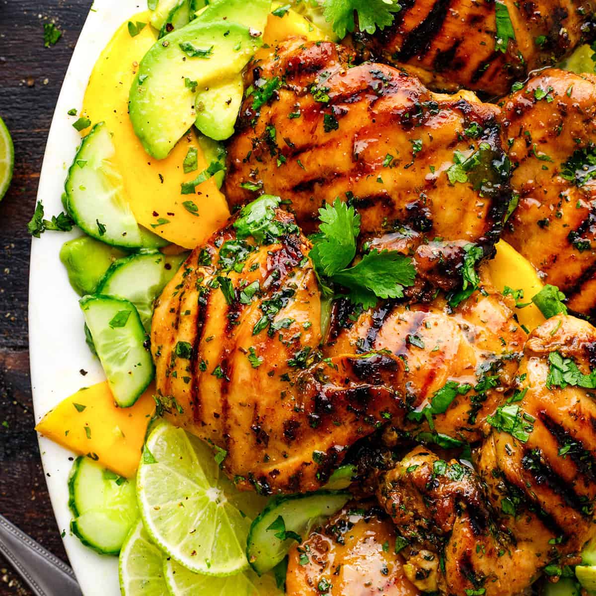 Cilantro Lime Chicken Grilled chicken thighs garnished with cilantro, served with sliced cucumbers, mango, and lime on a white plate.