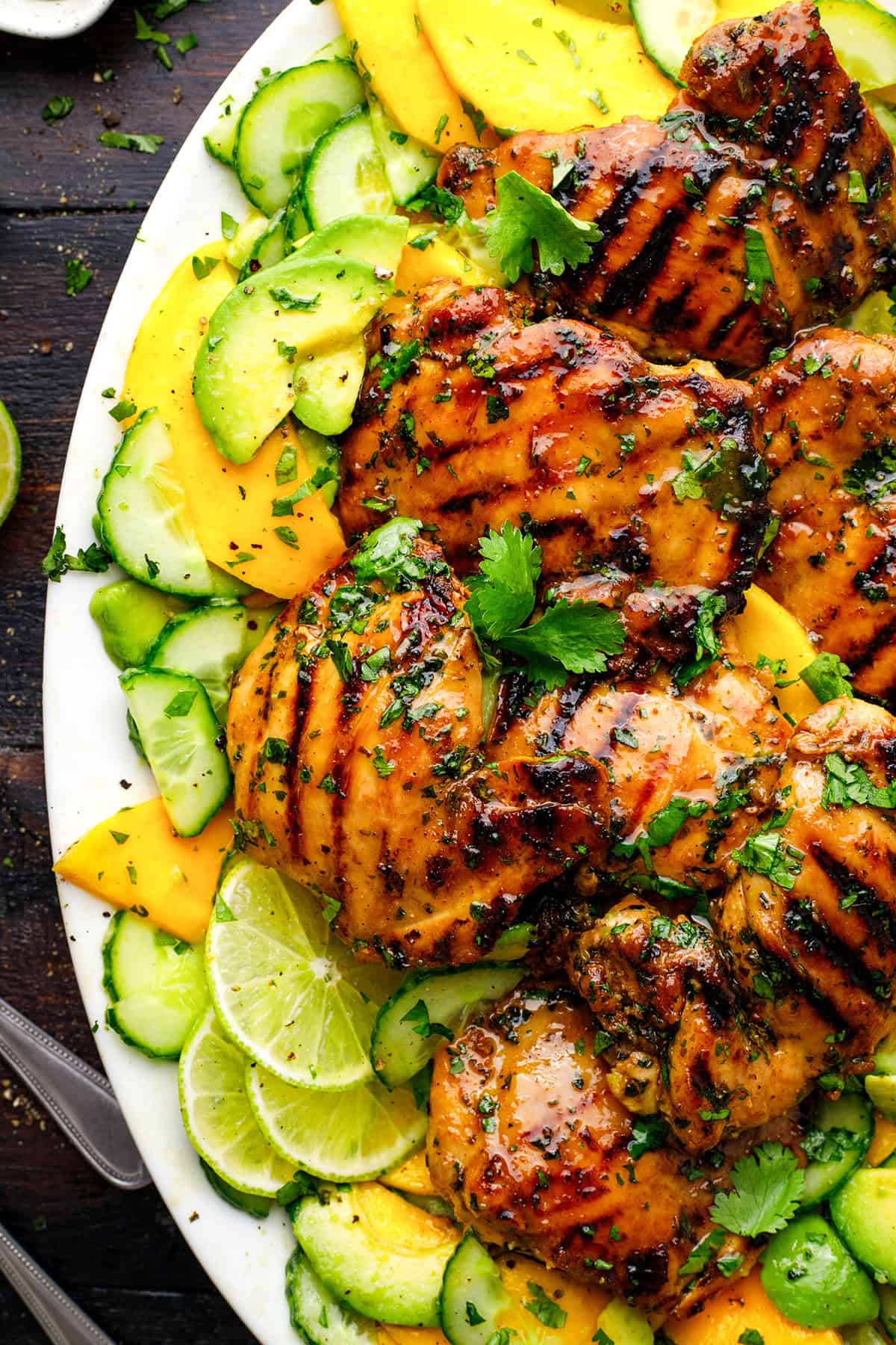Grilled Cilantro Lime Chicken Thighs Grilled glazed chicken thighs on a platter with sliced cucumbers, mango, lime, and fresh cilantro garnish.