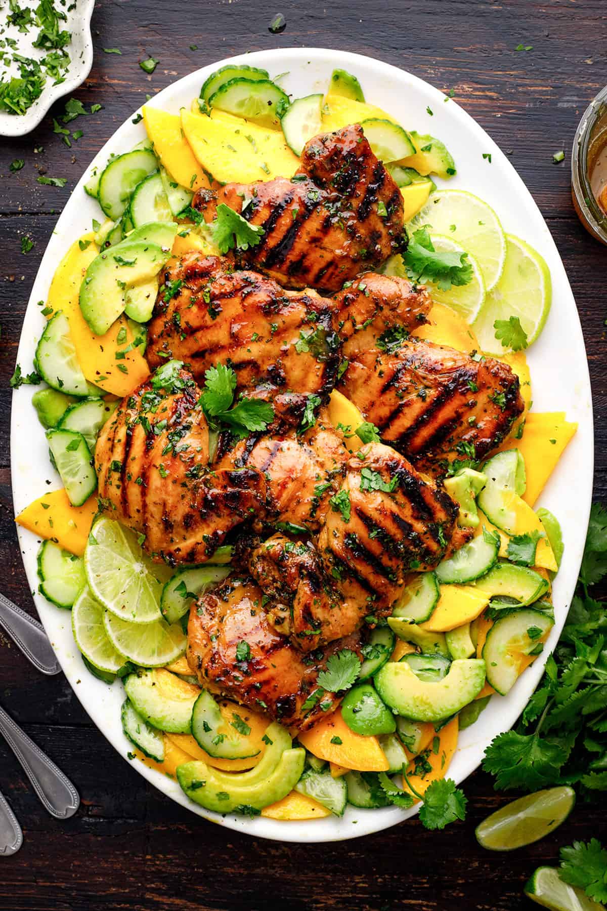 Grilled Cilantro Lime Chicken Thighs Grilled chicken thighs on a platter with sliced cucumbers, mango, avocado, lime wedges, and cilantro, arranged on a dark wooden table.