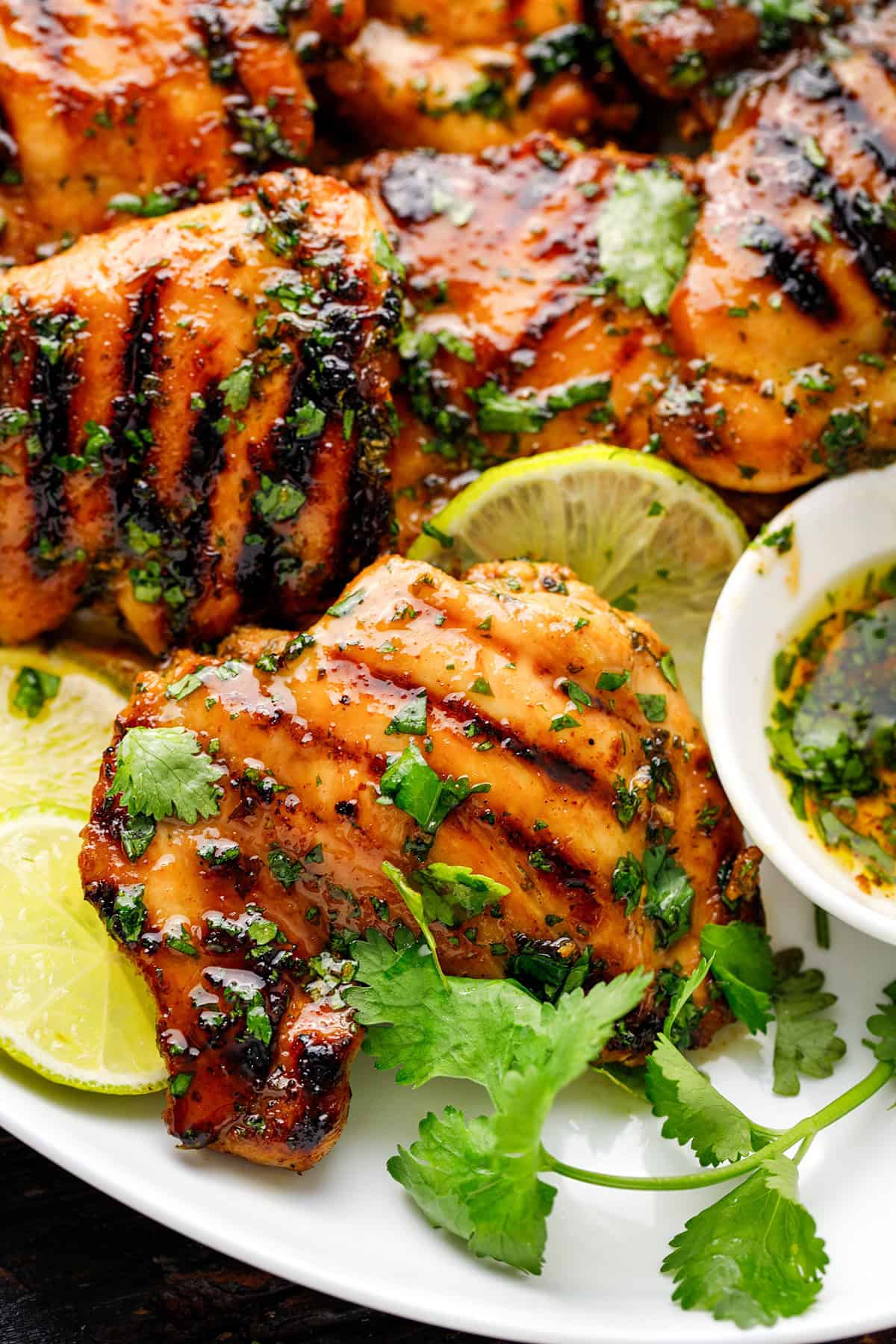Grilled Cilantro Lime Chicken Thighs Grilled chicken thighs garnished with fresh cilantro and lime slices, served on a white plate with a small bowl of sauce.