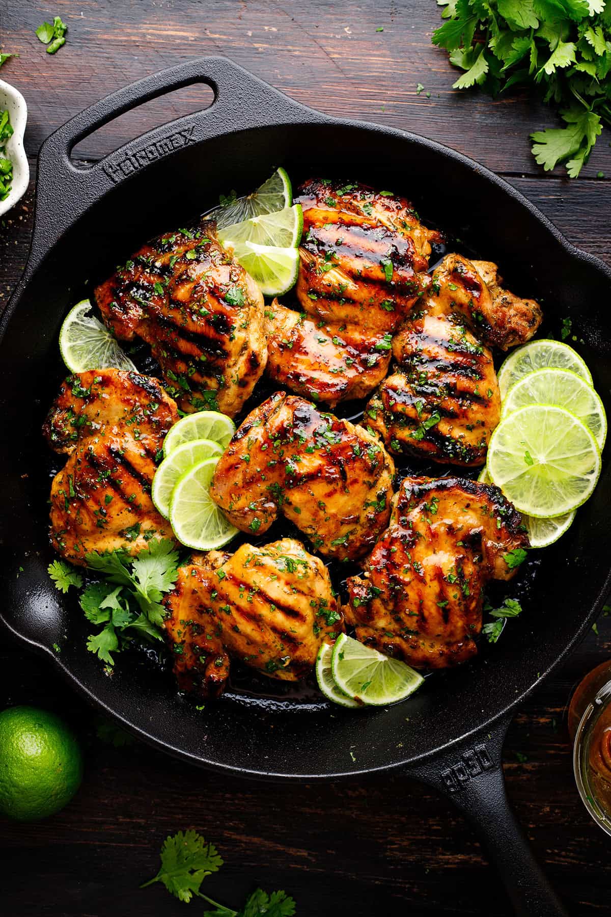Grilled Cilantro Lime Chicken Thighs Grilled chicken thighs in a cast iron skillet, garnished with fresh cilantro and lime slices, set on a dark wooden table.