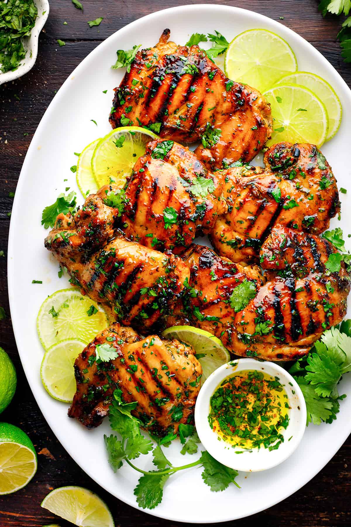 Cilantro-Lime Chicken Thighs Grilled chicken thighs garnished with chopped herbs on a white plate, surrounded by lime slices and a small bowl of herb sauce.