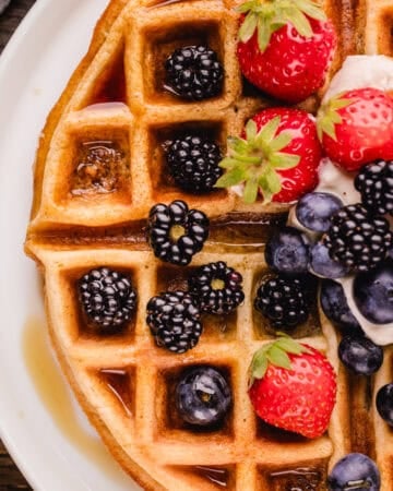 A waffle topped with fresh strawberries, blackberries, blueberries, cream, and syrup on a white plate.
