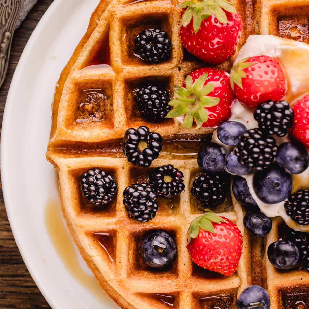 A waffle topped with fresh strawberries, blackberries, blueberries, cream, and syrup on a white plate.