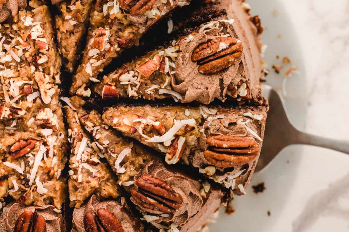 German Chocolate Cake A close-up of a sliced German chocolate cake topped with shredded coconut, pecans, and chocolate frosting, with a cake server lifting one slice on a white plate.