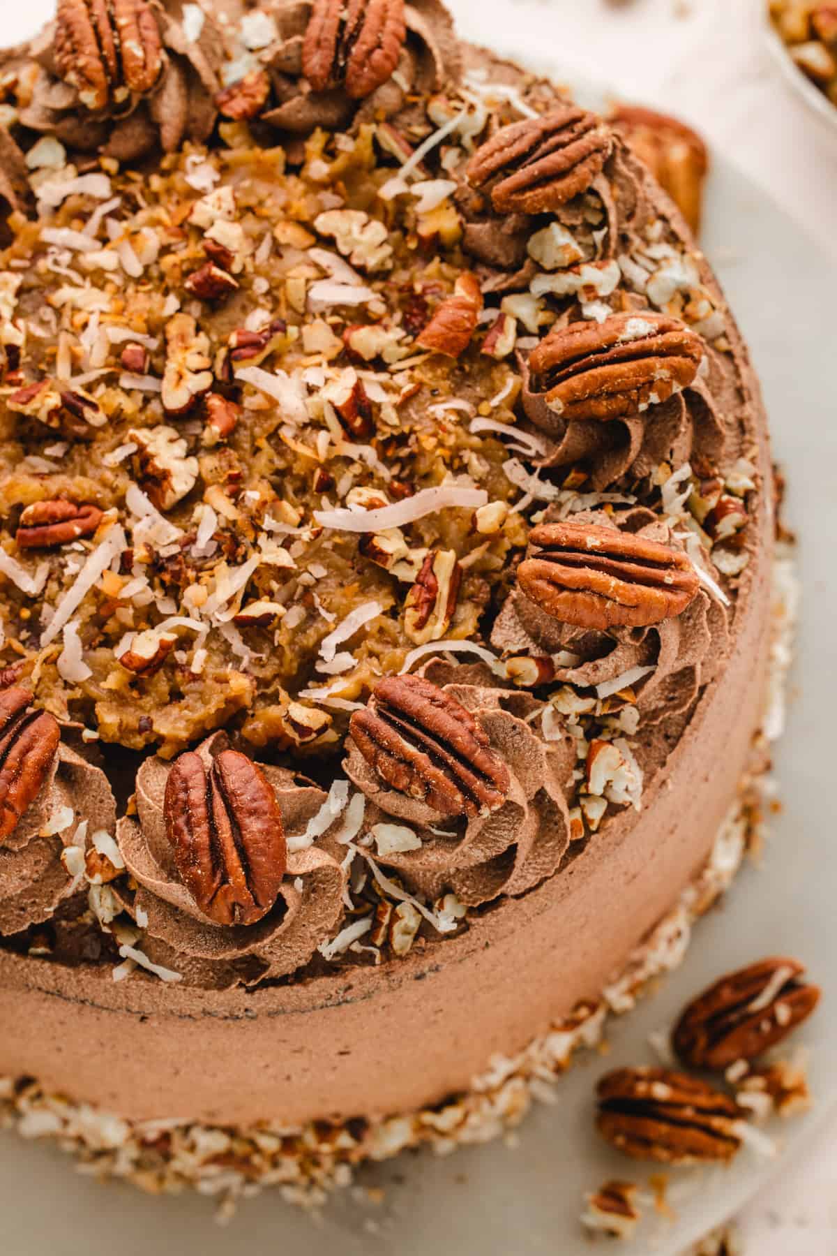 German Chocolate Cake A close-up of a German chocolate cake topped with chocolate frosting, pecans, shredded coconut, and chopped nuts on a white surface.