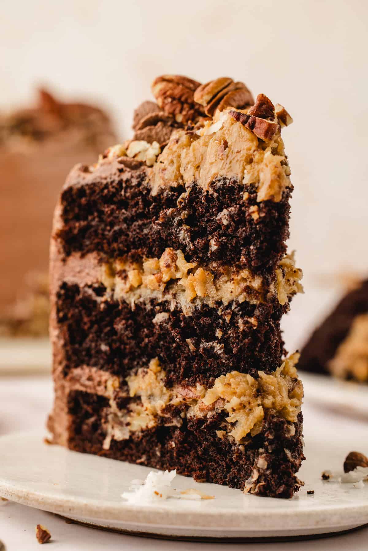 German Chocolate Cake A slice of chocolate layer cake with creamy filling and pecan pieces on top, served on a white plate.