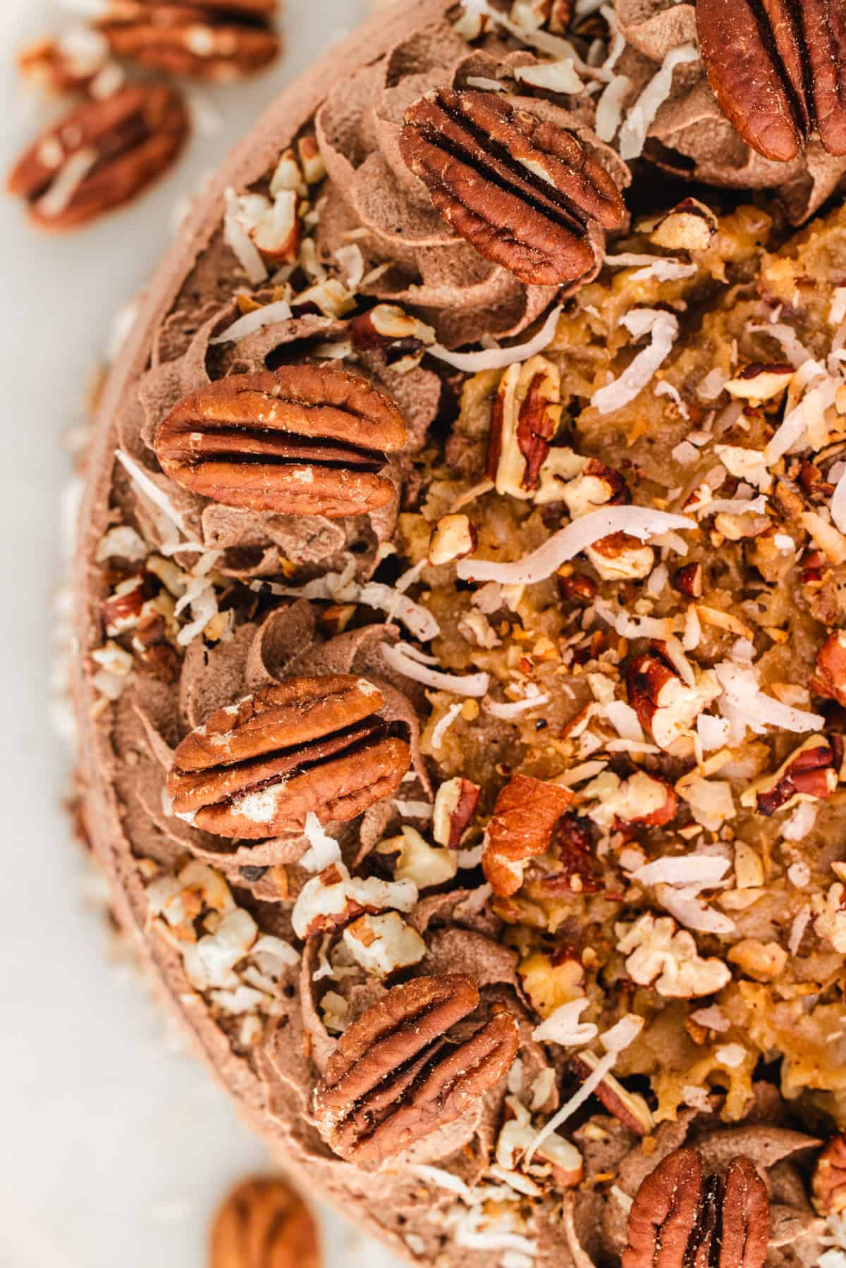 German Chocolate Cake Top view of a German chocolate cake topped with pecan halves, shredded coconut, and chocolate frosting, with some chopped pecans and coconut sprinkled on top and around the cake.