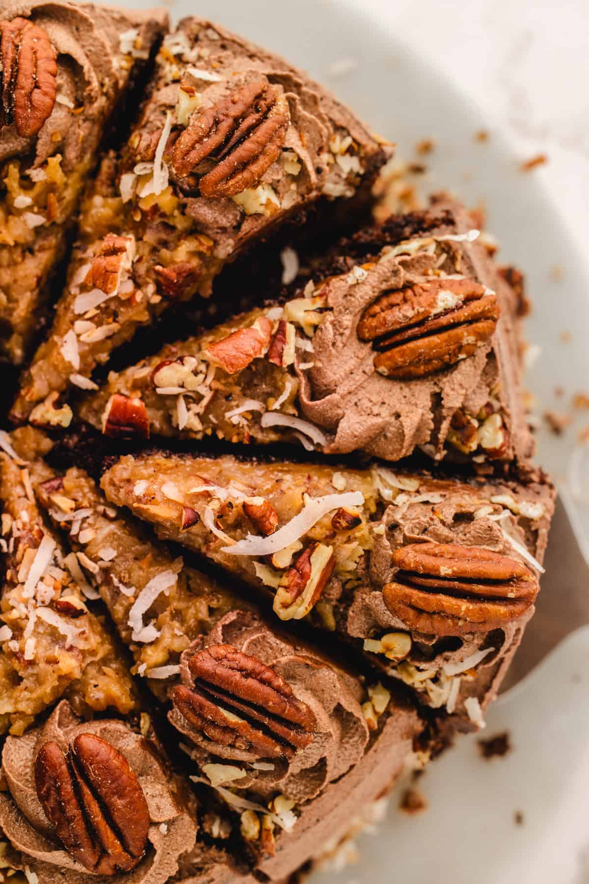 German Chocolate Cake Close-up of sliced German chocolate cake topped with pecans, shredded coconut, and chocolate frosting, arranged on a white plate.
