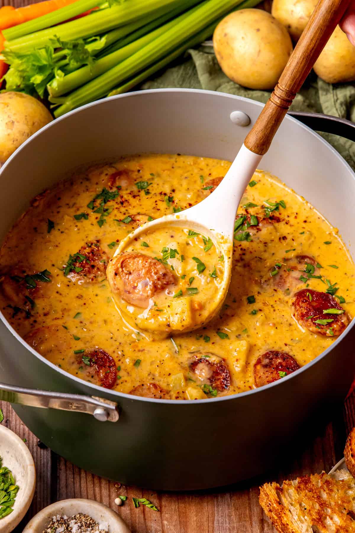 Kielbasa Potato Soup A ladle lifts cheesy sausage and potato soup from a pot, surrounded by potatoes, celery, fresh herbs, bread, and seasonings on a wooden table.
