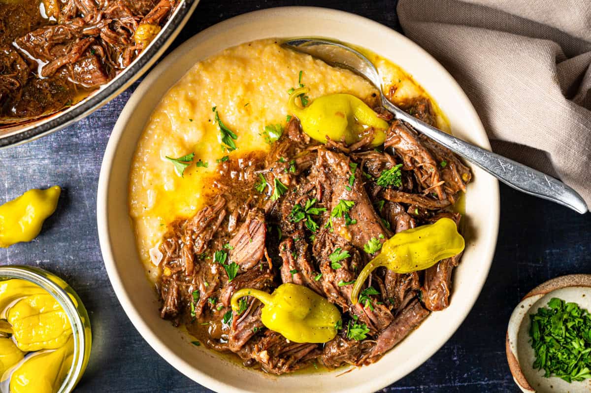 A bowl of shredded beef in gravy, garnished with pepperoncini peppers and chopped parsley, served over polenta.
