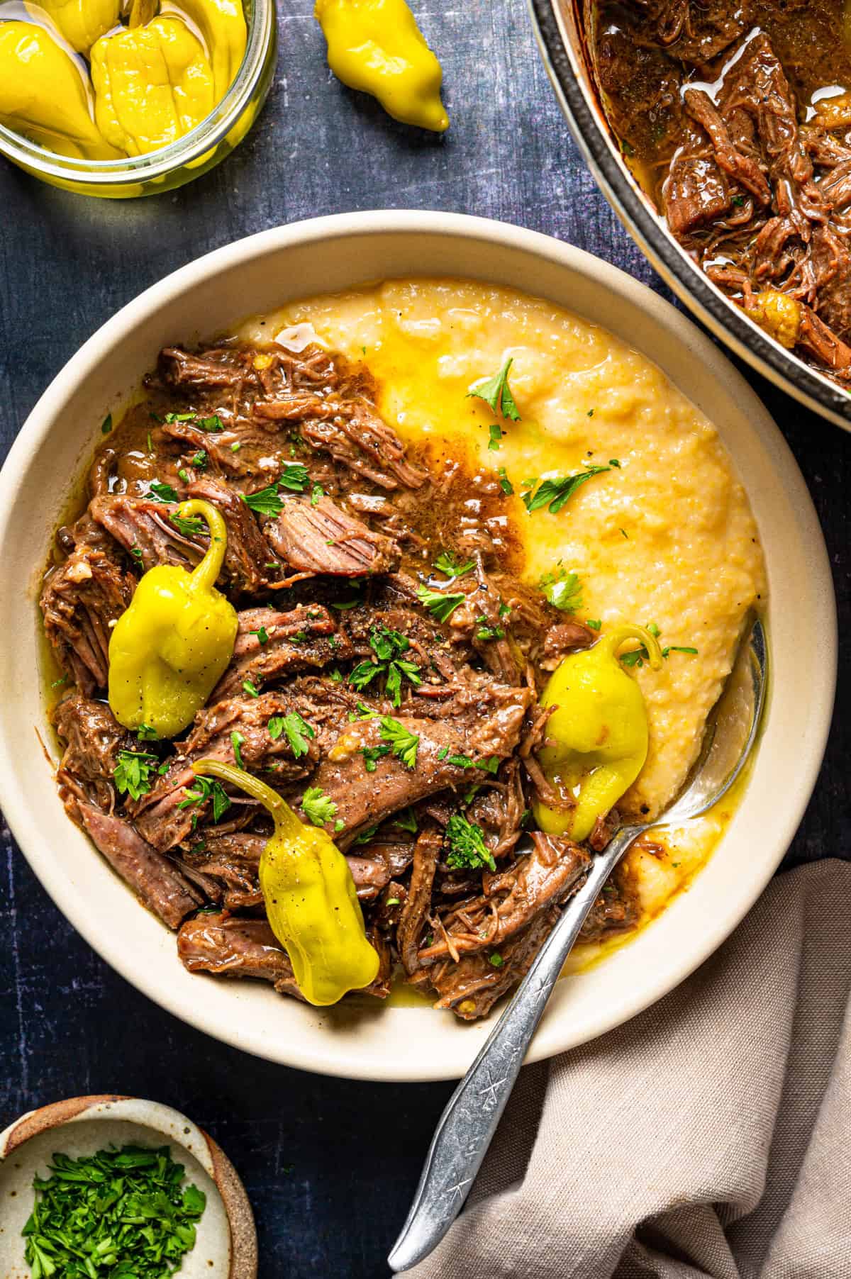 Mississippi Pot Roast (Easy Oven-Braised Chuck Roast) A bowl of shredded beef with yellow pepperoncini and chopped herbs served over creamy polenta.