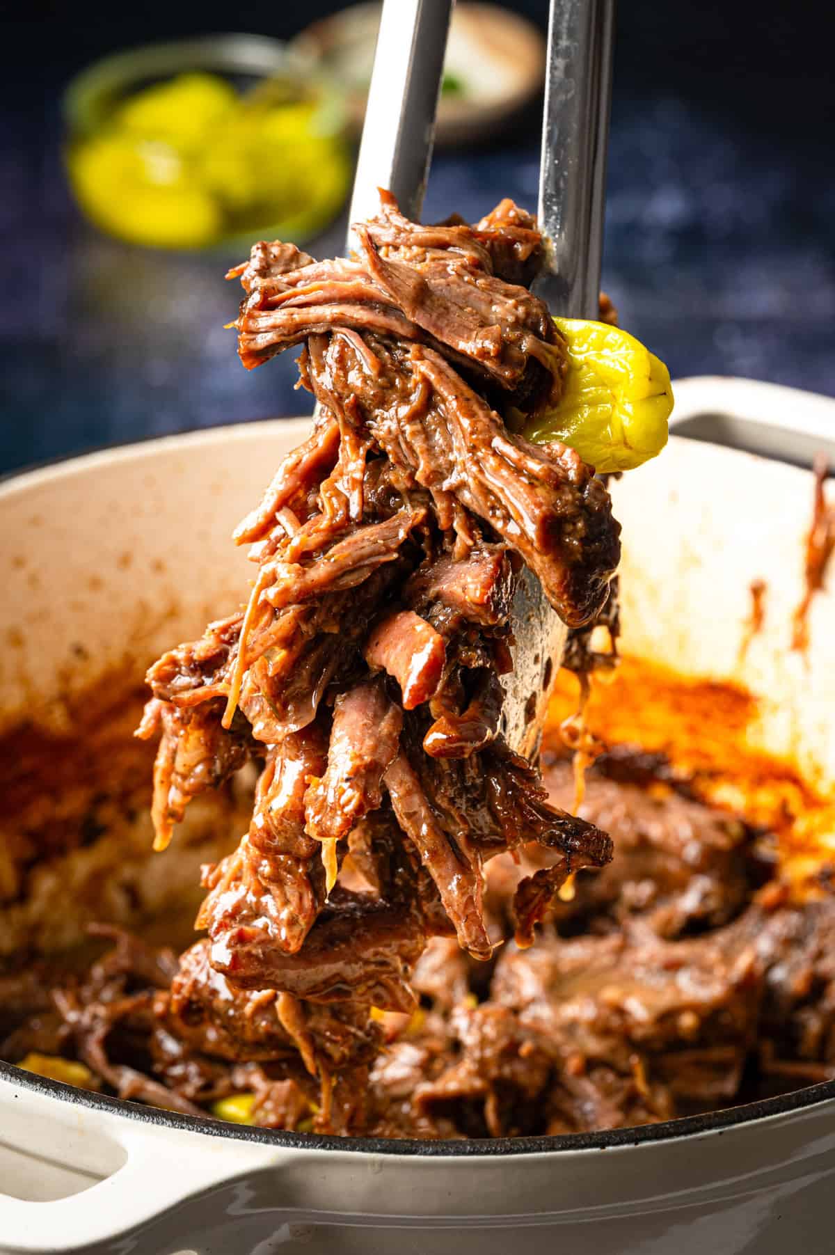 Mississippi Pot Roast (Easy Oven-Braised Chuck Roast) Tongs lifting shredded, tender beef from a pot, with visible spices and a yellow pepper, showcasing a cooked, saucy meat dish.