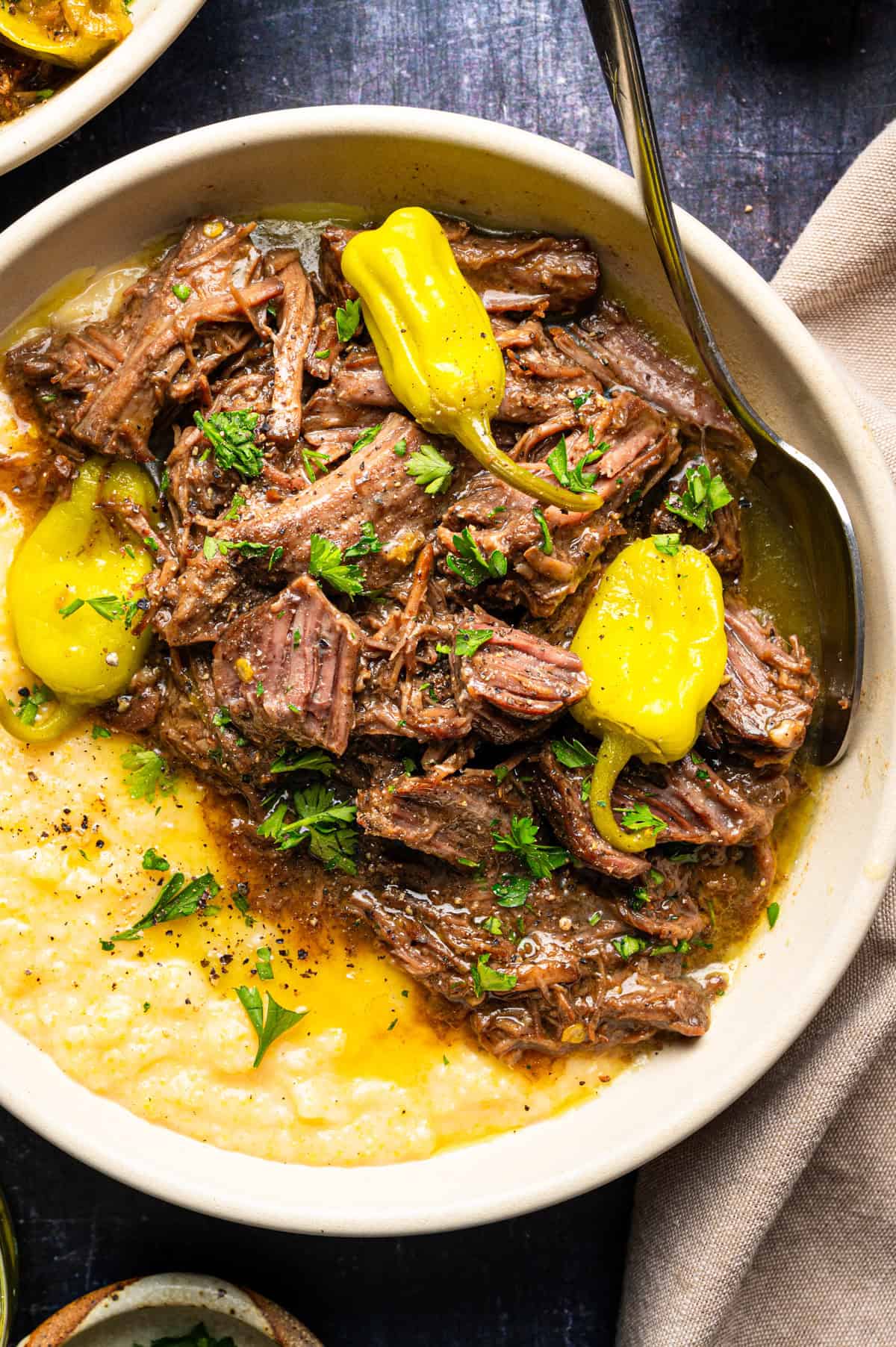Mississippi Pot Roast (Easy Oven-Braised Chuck Roast) A bowl of shredded roast beef with gravy, garnished with yellow peppers and fresh herbs, served over polenta.