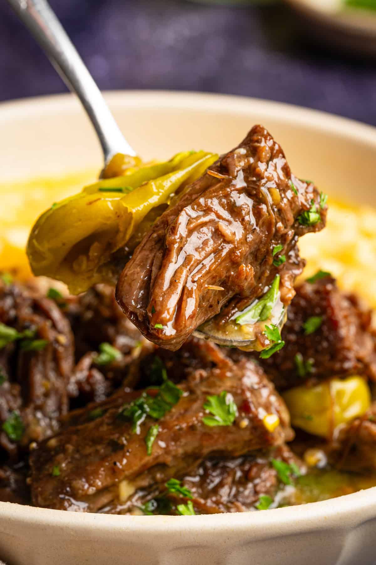 Mississippi Pot Roast (Easy Oven-Braised Chuck Roast) A close-up of a fork holding a tender piece of braised beef on top of creamy sauce, garnished with herbs, with more beef and sauce in the bowl below.
